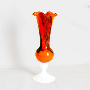 Puede incluir: Un jarrón de cristal naranja y rojo vintage con borde con volantes. El jarrón tiene una sección media bulbosa y se asienta sobre una base de pedestal blanca. El cristal tiene un efecto veteado con vetas oscuras.