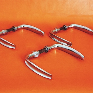 Op de afbeelding: Drie chromen deurgrepen op een oranje oppervlak. Elke handgreep heeft een gebogen ontwerp met een centraal verbindingsstuk. De handgrepen zijn reflecterend en tonen de omgeving. De algehele esthetiek is modern en strak.