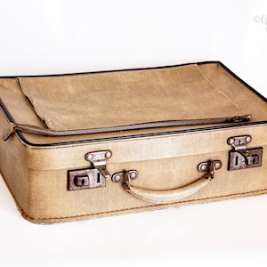 May include: A rectangular beige suitcase with a textured surface, featuring a dark brown trim, a central handle, and two metal latches. A horizontal zip runs across the top lid, and the case has a structured, boxy shape with visible stitching.