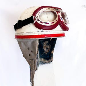 May include: A vintage-style white motorcycle helmet featuring a bright red horizontal stripe around the base. The helmet is equipped with dark maroon goggles that have silver frames and a black elastic strap. The interior lining is dark and worn.
