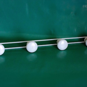 May include: A white metal coat rack for wall mounting, featuring four white, round knobs. The rack is set against a green backdrop. The knobs are spaced out along a horizontal bar.