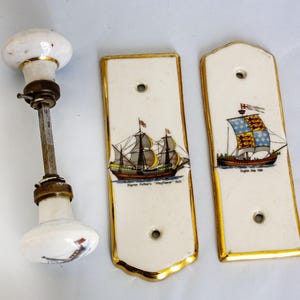 Può includere: Un pomello per porta vintage e due placche posteriori in porcellana. Il pomello è bianco con finiture dorate e un'asta di metallo. Le placche posteriori sono rettangolari con finiture dorate e presentano illustrazioni di navi con il testo: "Pilgrim Fathers 'Mayflower' 1620" e "English Ship 1588".