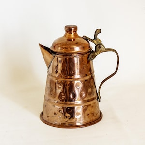 Puede incluir: Una tetera de cobre vintage con tapa y boquilla. La tetera tiene una textura martillada y un asa de latón. El cobre tiene un color marrón rojizo cálido. La tetera probablemente se usa para preparar té o café.
