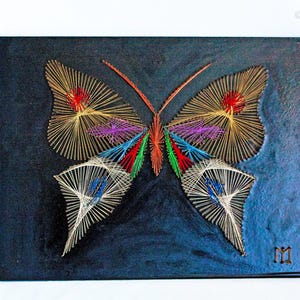 Pode incluir: Um design de borboleta em arte de fios sobre um fundo azul escuro texturizado. A borboleta apresenta padrões complexos criados com fios dourados, vermelhos, roxos, azuis, verdes e brancos. O design inclui asas simétricas e duas antenas.