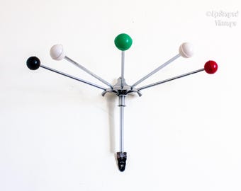 Può includere: Un attaccapanni vintage con finitura cromata e sei pomelli colorati. I pomelli sono neri, bianchi, verdi e rossi. L'attaccapanni è montato su una parete bianca e ha un gambo centrale.
