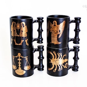 May include: Four matte black ceramic mugs with distinct gold zodiac illustrations. The mugs feature unique handles with a stacked, segmented design. The graphics depict Gemini, Libra, Virgo, and Scorpio signs in a detailed, metallic gold ink finish.