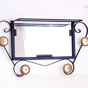 May include: Black metal wall mirror with a rectangular mirror and four gold-coloured knobs. The mirror has a shelf above it and decorative scrollwork on the sides. The text "UpStaged Vintages" is in the upper right corner.
