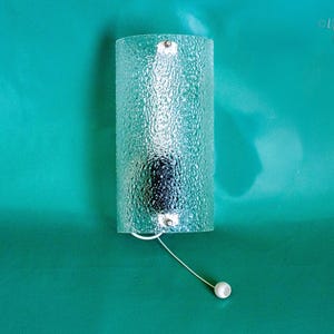 Puede incluir: Aplique de pared de vidrio transparente texturizado, de forma cilíndrica. El aplique tiene una cadena de tracción con una bola blanca en el extremo. El fondo es de color verde azulado.