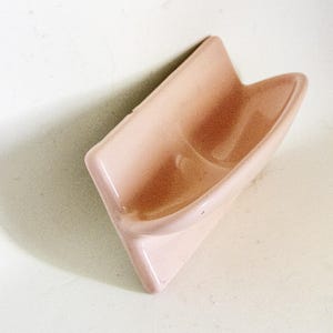 May include: A pale pink ceramic soap dish with a curved design. The dish is mounted on a wall.