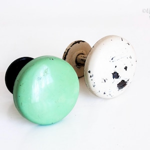 May include: Two round metal cabinet knobs. One knob is a smooth, solid mint green color with a black base. The second knob is off-white with visible black paint chips and wear, featuring a matching off-white base. Both knobs have a rounded top.