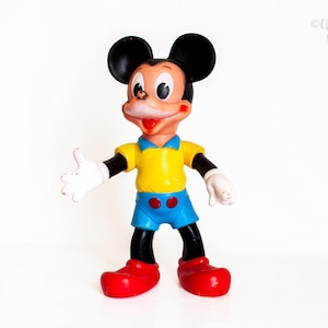 Puede incluir: Una figura vintage de Mickey Mouse con cabeza negra y grandes orejas redondas, cara color melocotón, camisa amarilla con cuello azul, pantalones cortos azules con dos botones rojos, guantes blancos y zapatos rojos grandes en pose acogedora.