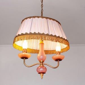 May include: Vintage chandelier featuring a white pleated lampshade with gold trim and fringe. The chandelier has three lights with peach-coloured accents and a gold-toned frame. A classic lighting fixture.