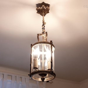 Puede incluir: Un candelabro antiguo de latón y vidrio cuelga del techo. La lámpara tiene una pantalla cilíndrica de vidrio, adornos de metal y tres luces interiores. El techo es blanco y la lámpara está iluminada.