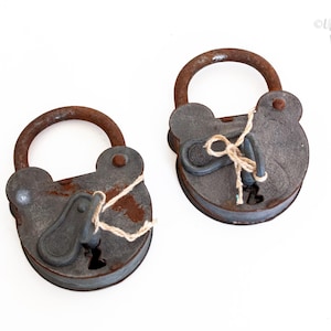 May include: Two weathered gray metal padlocks with rusted iron shackles. Each padlock features a key inserted into the keyhole, secured with a piece of thin twine tied around the shackle and the key. The metal surfaces show signs of oxidation.