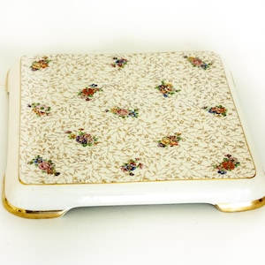 Può includere: Un sottopentola quadrato in ceramica bianca con un motivo floreale e un bordo dorato. Il sottopentola ha un bordo rialzato e quattro piedini.