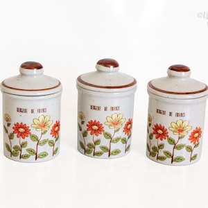 Può includere: Tre barattoli in ceramica con coperchi. I barattoli sono beige chiaro con bordo e coperchio marroni. Ogni barattolo presenta un motivo floreale con fiori rossi e gialli e la scritta "BERGER DE FRANCE".
