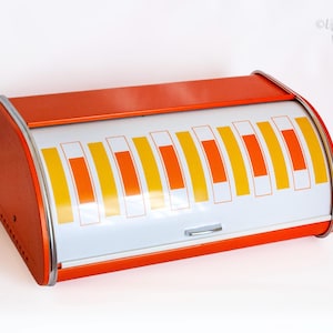 May include: A rectangular metal bread box with a roll-top lid. The frame is orange, while the curved sliding door is white, featuring a repeating pattern of vertical orange and yellow rectangular bars. A small silver handle is centered.