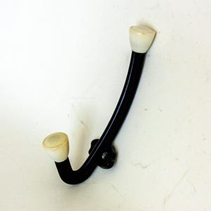 Pode incluir: Um gancho de casaco de metal preto com duas maçanetas de plástico branco. O gancho é curvo e tem um design simples.