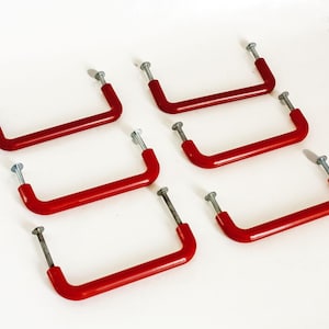 May include: Six red, U-shaped cabinet pulls with silver screws are arranged on a white background. The pulls are a vibrant red colour and appear to be made of metal. They are likely for furniture.