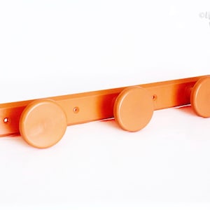 May include: An orange wall-mounted coat rack with three round knobs. The rack is made of plastic and has a rectangular base with mounting holes. The knobs are evenly spaced along the rack, providing a simple and functional design.