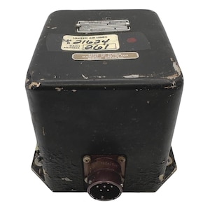 May include: A black, rectangular industrial control unit with a metallic connector port at the base. The top features several identification plates, including one labeled United Air Lines with handwritten numbers 21624 and 261, plus technical text.