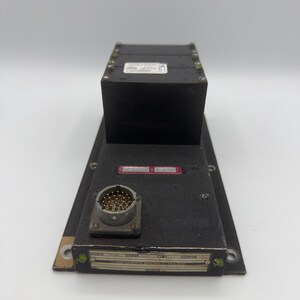 May include: A black rectangular electronic control unit featuring a raised top section, a circular multi-pin connector, and a metallic identification plate labeled Barber-Colman Co. Rockford, Ill. U.S.A. with a red serial number tag attached.