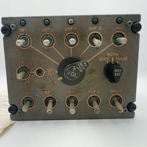 May include: A vintage gray metal aircraft audio control panel featuring multiple toggle switches, a central volume knob labeled PULL FOR PA, and a secondary knob labeled BOTH VOICE RANGE NAV ADF. The panel includes labels for INT, VHF-1, VHF-2, VHF-3, PA, OXY, PTT, SERV INT, OFF, NAV-1, ADF-1, MKR, ADF-2, and NAV-2.