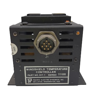 May include: A black metal windshield temperature controller by Pacific Electro Dynamics, Inc. The unit features a circular J1 connector, ventilation slots, and a label reading: WINDSHIELD TEMPERATURE CONTROLLER, PART NO. 617-1, SERNO 1102.