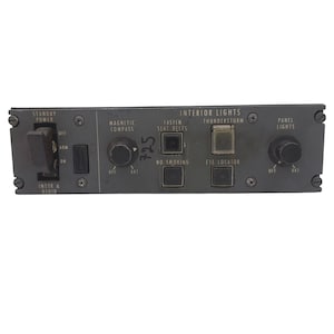 May include: A dark gray aircraft control panel featuring a Standby Power switch, knobs for Magnetic Compass and Panel Lights, and push-button switches for Fasten Seat Belts, No Smoking, Thunderstorm, and Eye Locator, with the text Interior Lights.