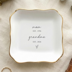 Puede incluir: Una bandeja de cerámica blanca con borde dorado, inscrita con las palabras "mamá" y "abuela" en escritura negra, junto con los años 1996 y 2026. La bandeja está rodeada de joyas y un pequeño libro encuadernado en cuero.