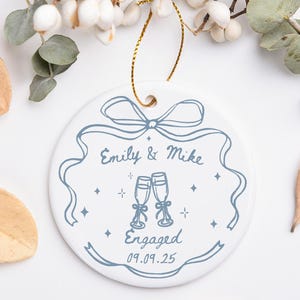 May include: White ceramic ornament with a blue ribbon design and the text "Emily & Mike Engaged 09.09.25". The ornament features two champagne glasses and star accents, with a gold string for hanging. Decorative leaves and berries surround the ornament.