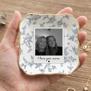 Peut inclure: Un plat décoratif en céramique avec un motif floral blanc et bleu, bordé d'or. Le plat contient une petite photo avec le texte "I love you mom". Des bijoux en or, dont un collier et des boucles d'oreilles, sont également visibles.