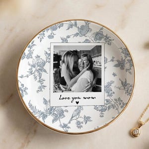 Könnte beinhalten: Ein dekorativer Teller mit weißen und blauen Blumenmustern und einem goldenen Rand, der ein Schwarz-Weiß-Foto mit dem Text "Love you mom" enthält. Eine goldene Halskette mit Anhänger und Perlenohrringe liegen neben dem Teller.
