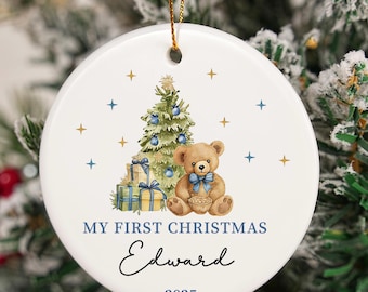 May include: White ceramic Christmas ornament with a teddy bear, Christmas tree, and gifts design. The text reads "My First Christmas Edward 2025". The ornament is round and has a gold string for hanging.