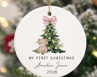 Peut inclure: Une décoration ronde en céramique blanche ornée d'une illustration aquarelle d'un sapin de Noël vert surmonté d'un nœud vichy rose et d'un petit ours en peluche avec un nœud rose. Le texte indique MY FIRST CHRISTMAS Amalia Jean 2026.