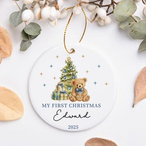 May include: A white ceramic Christmas ornament with a gold string. The ornament features a Christmas tree, a teddy bear, and the text "MY FIRST CHRISTMAS Edward 2025". The design includes blue and gold accents.