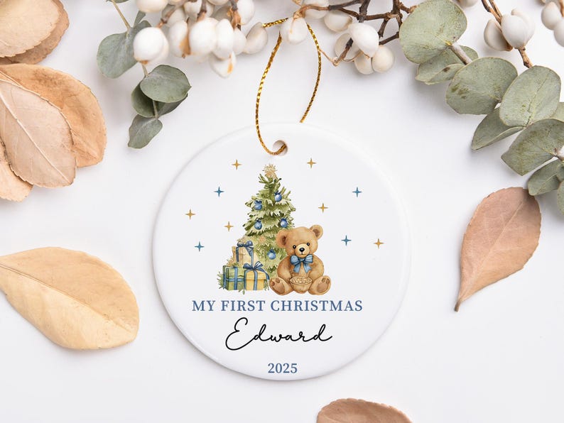 May include: A white ceramic Christmas ornament with a gold string. The ornament features a Christmas tree, a teddy bear, and the text "MY FIRST CHRISTMAS Edward 2025". The design includes blue and gold accents.