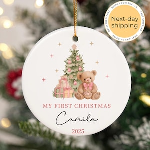 May include: Round white Christmas ornament featuring a festive design. It includes a Christmas tree, teddy bear, and gifts. The text reads "My First Christmas", the name "Camila", and the year "2025". A gold string is attached.