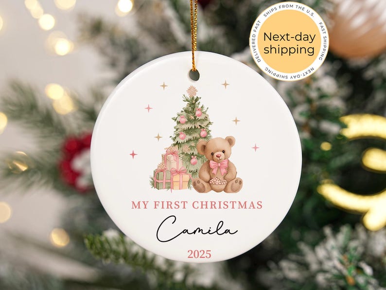 May include: Round white Christmas ornament featuring a festive design. It includes a Christmas tree, teddy bear, and gifts. The text reads "My First Christmas", the name "Camila", and the year "2025". A gold string is attached.