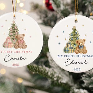 May include: Two white, round Christmas ornaments with gold string. Each features a teddy bear, Christmas tree, and gifts. One says "My First Christmas Camila 2025" and the other says "My First Christmas Edward 2025".