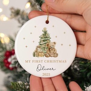 May include: A round white ceramic Christmas ornament with a gold string. It features a watercolour illustration of a Christmas tree, teddy bear, and gifts. Text reads "MY FIRST CHRISTMAS" and a name and year.