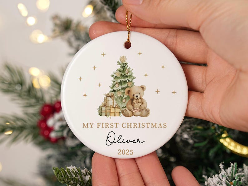 May include: A round white ceramic Christmas ornament with a gold string. It features a watercolour illustration of a Christmas tree, teddy bear, and gifts. Text reads "MY FIRST CHRISTMAS" and a name and year.