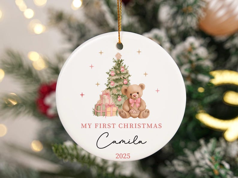 May include: A white ceramic Christmas bauble with a watercolour design. It features a Christmas tree, a teddy bear, and wrapped gifts. The text reads "My First Christmas" and "Camila 2025". A gold string is attached for hanging.