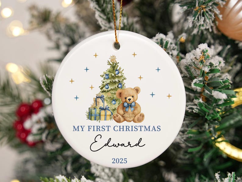 May include: Round white ceramic Christmas ornament featuring a teddy bear, Christmas tree, and gifts. Text reads "My First Christmas Edward 2025". The ornament has a gold string for hanging, perfect for a festive tree.