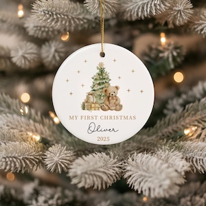 May include: A white ceramic Christmas bauble with a gold string. The bauble features a Christmas tree, teddy bear, and gifts. Text reads "MY FIRST CHRISTMAS" and "Oliver 2025". The bauble is hanging on a Christmas tree.