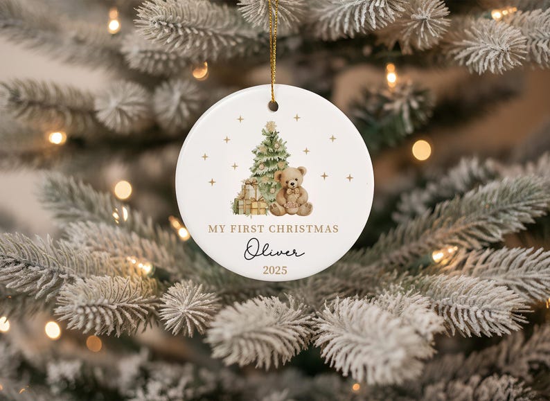 May include: A white ceramic Christmas bauble with a gold string. The bauble features a Christmas tree, teddy bear, and gifts. Text reads "MY FIRST CHRISTMAS" and "Oliver 2025". The bauble is hanging on a Christmas tree.
