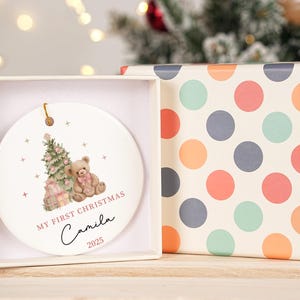 May include: A white ceramic Christmas bauble featuring a teddy bear, Christmas tree, and gifts design. The bauble is in a white box next to a box with a colourful polka dot pattern. Text on the bauble reads "MY FIRST CHRISTMAS Camila 2025".