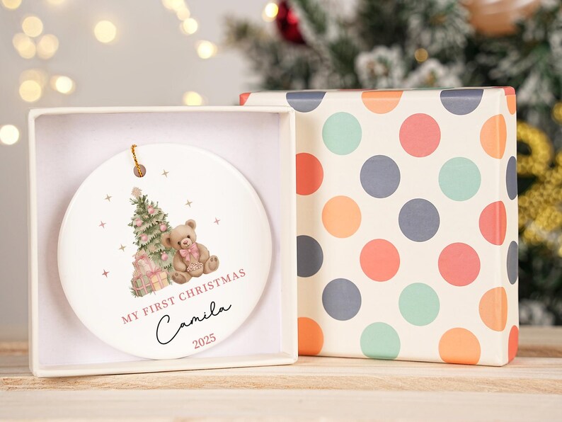 May include: A white ceramic Christmas bauble featuring a teddy bear, Christmas tree, and gifts design. The bauble is in a white box next to a box with a colourful polka dot pattern. Text on the bauble reads "MY FIRST CHRISTMAS Camila 2025".