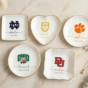 May include: White ceramic trinket dishes in various shapes, each with a gold rim and personalized text. Designs include a paw print, university logos, and names like Amelia, Sarah, Hannah, and Madison, with the text "Class of 2026".