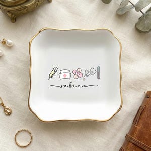 Può includere: Vassoio portagioie in ceramica bianca con bordo dorato, con icone mediche e il nome "sabina" in corsivo. Il vassoio è circondato da gioielli e un taccuino rilegato in pelle.